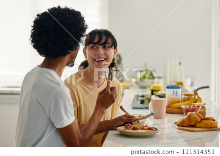 Young cheerful woman looking at her husband or boyfriend holding fork with tiny piece of homemade pizza by her mouth during breakfast 111314351