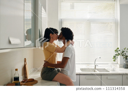 Young affectionate woman in casualwear sitting on kitchen counter and kissing her husband keeping his hands around her waist 111314358