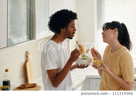 Side view of young multicultural couple with salad in bowls having breakfast and looking at one another in the kitchen Side view of young multicultural couple with salad in bowls having breakfast and looking at one another in the kitchen 111314368