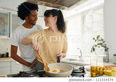 Pretty young woman with frying pan cooking scrambled eggs for breakfast while standing by electric stove and looking at her husband 111314374