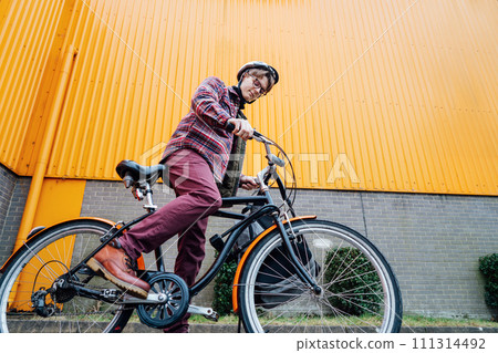 Bottom view Young stylish man in helmet riding on cruiser city Bicycle near orange Wall. Student on the way to college on bike. Eco friendly transportation for carbon footprint. Sustainable lifestyle Bottom view Young stylish man in helmet riding on cruiser city Bicycle near orange Wall. Student on the way to college on bike. Eco friendly transportation for carbon footprint. Sustainable lifestyle 111314492