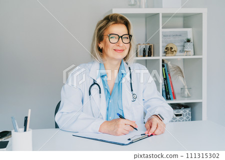 Confident mature middle aged woman doctor medical nurse, female physician practitioner in white coat with stethoscope sitting at working place at hospital office and making notes during appointment. 111315302