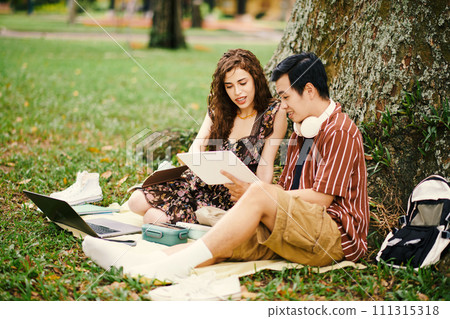 Two young intercultural students preparing presentation of new school project and discussing its points while sitting under tree in park Two young intercultural students preparing presentation of new school project and discussing its points while sitting under tree in park 111315318