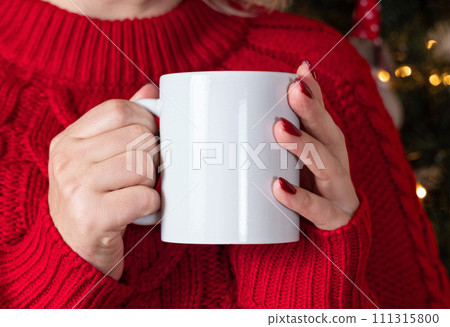 Woman in red sweater with white mug in hands against Christmas tree with lights, mock up Woman in red sweater with white mug in hands against Christmas tree with lights, mock up 111315800