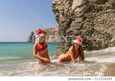 Women Santa hats ocean play. Seaside, beach daytime, enjoying beach fun. Two women in red swimsuits and Santa hats are enjoying themselves in the ocean waves. Women Santa hats ocean play. Seaside, beach daytime, enjoying beach fun. Two women in red swimsuits and Santa hats are enjoying themselves in the ocean waves. 111316285