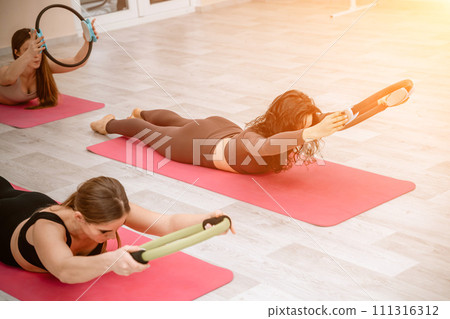 A group of six athletic women doing pilates or yoga on pink mats in front of a window in a beige loft studio interior. Teamwork, good mood and healthy lifestyle concept. 111316312