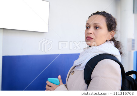 Close-up young woman commuter holding mobile phone and looking away, standing at information desk in the railway station Close-up young woman commuter holding mobile phone and looking away, standing at information desk in the railway station 111316708