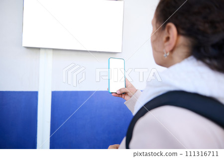 Focus on white blank digital mockup screen of a smartphone in the hand of a commuter taking photo of information desk Focus on white blank digital mockup screen of a smartphone in the hand of a commuter taking photo of information desk 111316711