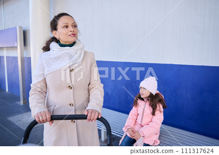 Young woman, mother pushing baby pram, standing near her daughter on the railway station platform, waiting for train 111317624