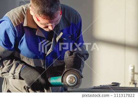 Man in overalls and safety glasses works with angle grinder in workshop. Worker grinds metal Man in overalls and safety glasses works with angle grinder in workshop. Worker grinds metal 111317822