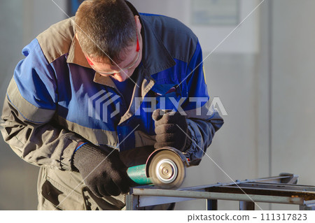 Man in overalls and safety glasses works with angle grinder in workshop. Worker grinds metal Man in overalls and safety glasses works with angle grinder in workshop. Worker grinds metal 111317823