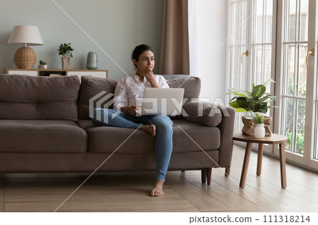 Indian woman sit on couch with laptop looks pensive Indian woman sit on couch with laptop looks pensive 111318214