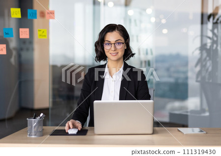 Businesswoman in business suit inside office working with laptop, woman smiling and looking at camera, female worker in business suit is satisfied with achievement results at workplace. 111319430