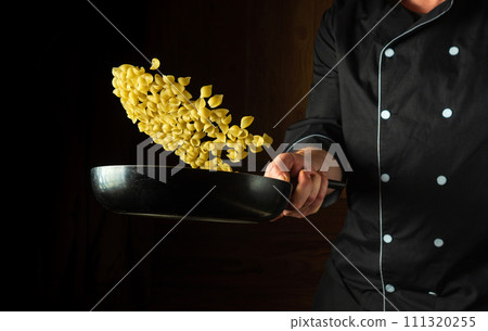 Flight of pasta over the frying pan in the hand of the cook. Concept of cooking shell type pasta in restaurant kitchen for lunch Flight of pasta over the frying pan in the hand of the cook. Concept of cooking shell type pasta in restaurant kitchen for lunch 111320255