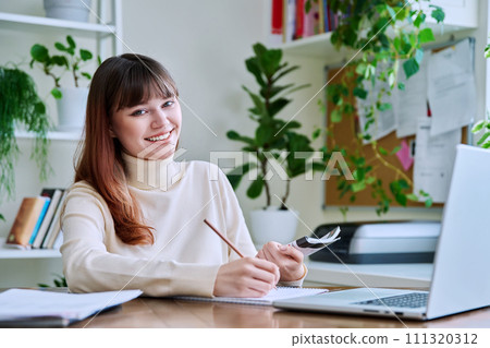 Young female college student studying at home using laptop, writing in notebook 111320312
