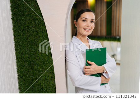 Portrait of positive young woman doctor or nurse in medical coat holding clipboard with with medical report standing posing in hallway, smiling looking at camera. Concept of healthcare. Portrait of positive young woman doctor or nurse in medical coat holding clipboard with with medical report standing posing in hallway, smiling looking at camera. Concept of healthcare. 111321342