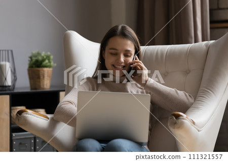 Young woman talks on smartphone seated on armchair with laptop 111321557