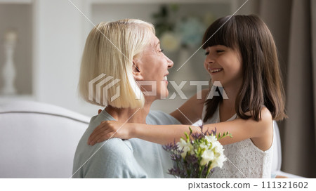 Smiling little granddaughter congratulating grandmother, presenting flowers 111321602