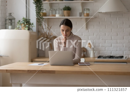 Serious woman sit at table in domestic kitchen with laptop Serious woman sit at table in domestic kitchen with laptop 111321857