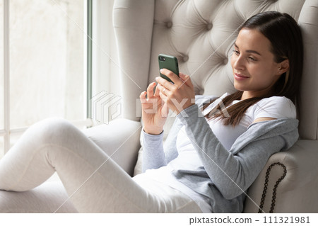 Satisfied young woman using phone and relaxing in cozy armchair Satisfied young woman using phone and relaxing in cozy armchair 111321981