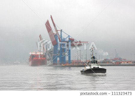Hamburg, Germany - December 25, 2012: Tugboat on the Elbe river in foggy weather. Silhouettes of a shipyard cranes in the port of Hamburg. 111323473