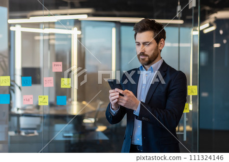 Serious businessman in suit using smartphone in modern office. Focused on finance, leadership, and corporate lifestyle. 111324146