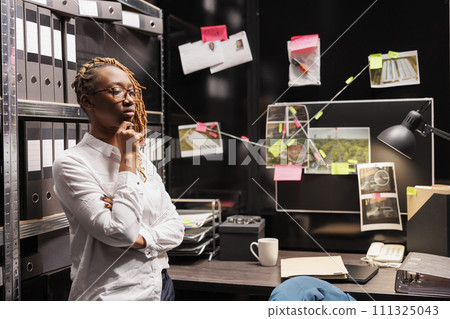 Pensive african american woman detective thinking about solving crime and rubbing chin. Thoughtful private investigator analyzing evidence while standing in agency office room 111325043