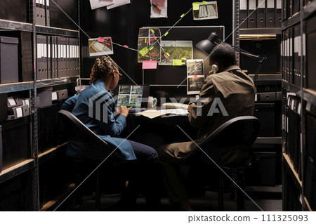Private investigators checking suspect photos on laptop and talking with client on landline phone. Detective partners conducting investigation together in agency dark office 111325093