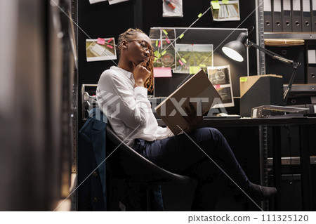 Thoughtful woman investigator reviewing forensic expertise report and rubbing chin in police office room. African american detective reading crime case file and analyzing information 111325120