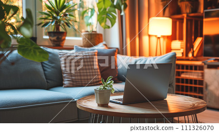 A home office nook warmly lit by a table lamp, with a laptop ready for use on a wooden round table, flanked by comfortable pillows and indoor greenery. 111326070