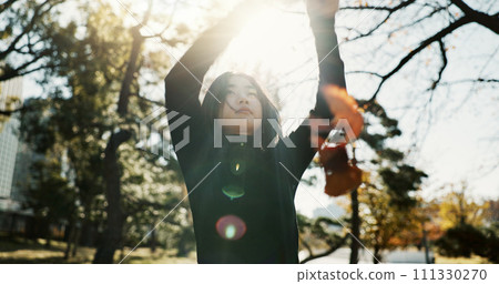 Playful, leaves, and woman with arms raised, sunshine and nature with lens flare, relaxing and peaceful. Person, outdoor and girl in autumn, fresh air and wellness with weekend break and freedom 111330270