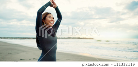 Surfer, Japanese and woman stretching at beach, happy for fitness and extreme sports with ocean view, waves and travel. Flexibility, warm up and exercise outdoor, surf and nature with smile in Japan 111332119
