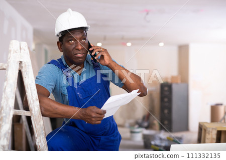 Worker in blue overalls communicates on a mobile phone in renovated room 111332135