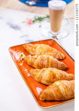 Four golden croissants on an orange plate with coffee cup and flowers on white marble table 111336775