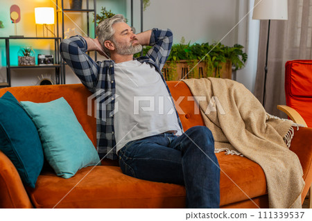 Tired senior middle-aged man stretching arms and resting relaxing on sofa in living room at home Tired senior middle-aged man stretching arms and resting relaxing on sofa in living room at home 111339537