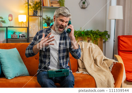 Sad disinterested senior man talking on vintage retro wired telephone with hotline helpline service 111339544