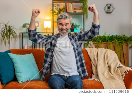 Happy Caucasian senior man watching TV shouting, celebrating success, winning, goal achievement Happy Caucasian senior man watching TV shouting, celebrating success, winning, goal achievement 111339616