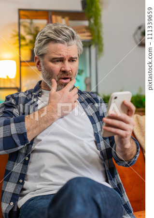 Shocked sad mature man holding smartphone reading negative message trouble feels annoyed at home Shocked sad mature man holding smartphone reading negative message trouble feels annoyed at home 111339659