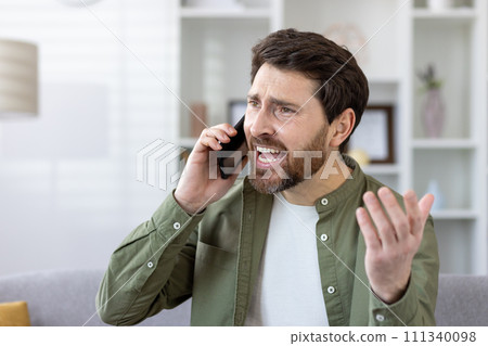 Focused mature bearded man in a casual shirt having a conversation on smartphone at his well-lit living room. 111340098