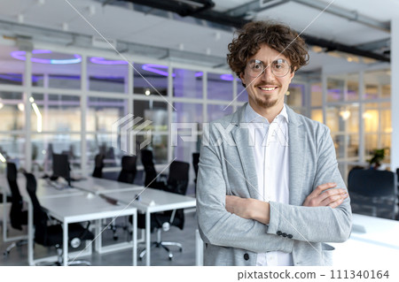 Portrait of a cheerful young businessman with curly hair confidently standing in a contemporary office space, arms crossed. Portrait of a cheerful young businessman with curly hair confidently standing in a contemporary office space, arms crossed. 111340164