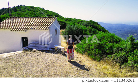 Corfu, Greece summer 2023. On the top of the mountain there is a small church a girl in a multi-colored skirt walks with flowers in her hands 111340261