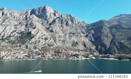 A drone flies over the Boka-Kotor Bay from one shore to the other. The houses of the city, which are located along the coast, are clearly visible. Cars drive on the roads. The sun illuminates the moun 111340283