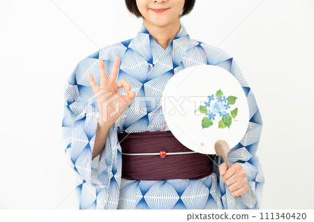 A middle-aged woman wearing a blue yukata and holding a fan and making a circle sign with her fingers 111340420
