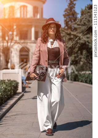 Woman park city. Stylish woman in a hat walks in a park in the city. Dressed in white corset trousers and a pink jacket with a bag in her hands. Woman park city. Stylish woman in a hat walks in a park in the city. Dressed in white corset trousers and a pink jacket with a bag in her hands. 111343123