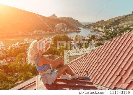 Woman sits on rooftop, enjoys town view and sea mountains. Peaceful rooftop relaxation. Below her, there is a town with several boats visible in the water. Rooftop vantage point. 111343139