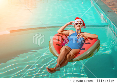 A happy woman in a blue bikini, a red and white Santa hat and sunglasses poses in the pool in an inflatable circle with a watermelon pattern, holding a glass of champagne in her hands. Christmas A happy woman in a blue bikini, a red and white Santa hat and sunglasses poses in the pool in an inflatable circle with a watermelon pattern, holding a glass of champagne in her hands. Christmas 111343146