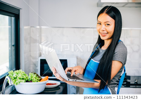 In the kitchen a smiling woman stands enjoying cooking and using a laptop for tutorials. Emphasizing the joy and convenience of technology in meal preparation and exploration. 111343423