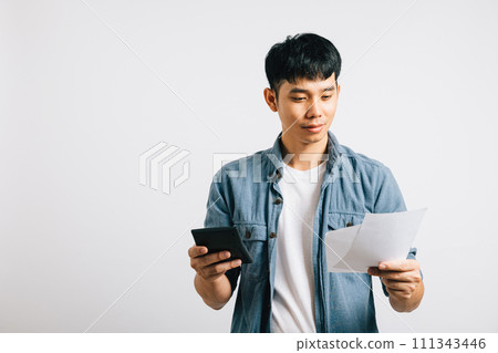 Young man in an isolated studio shot, holding a paper and calculator, displaying a sad, tired, and strained look while calculating bills. His worry over the financial problem is clear. Young man in an isolated studio shot, holding a paper and calculator, displaying a sad, tired, and strained look while calculating bills. His worry over the financial problem is clear. 111343446