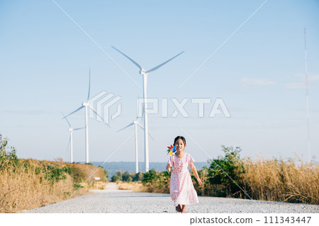 Little girl's playful learning near windmills holding pinwheels with joy. Embracing wind energy education in a cheerful clean electricity generating wind turbine landscape. 111343447