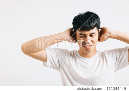 An Asian man in pain, covering his ears with his palms and closing his eyes due to loud noise. Studio shot isolated on white background, conveying his distress and the need for quiet. An Asian man in pain, covering his ears with his palms and closing his eyes due to loud noise. Studio shot isolated on white background, conveying his distress and the need for quiet. 111343449